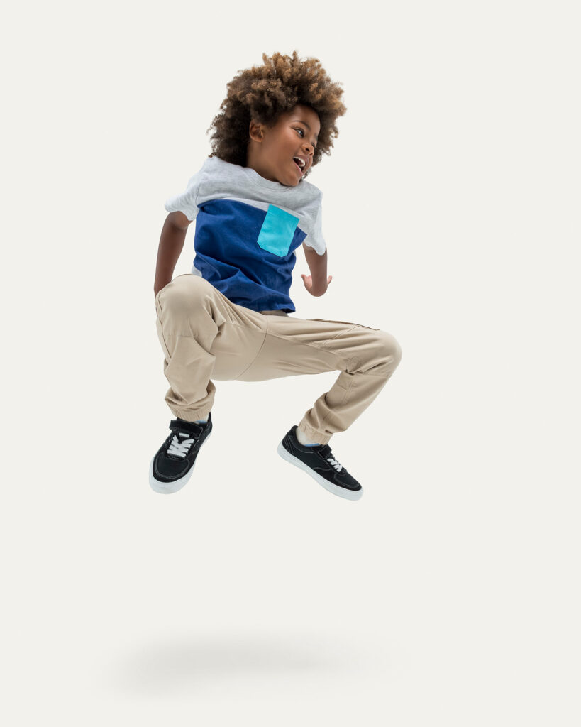 An energetic young boy with an afro hairstyle caught mid-air while jumping excitedly. He is wearing a grey and blue color-blocked T-shirt with a bright turquoise pocket, khaki jogger pants, and black sneakers against a solid light grey background.