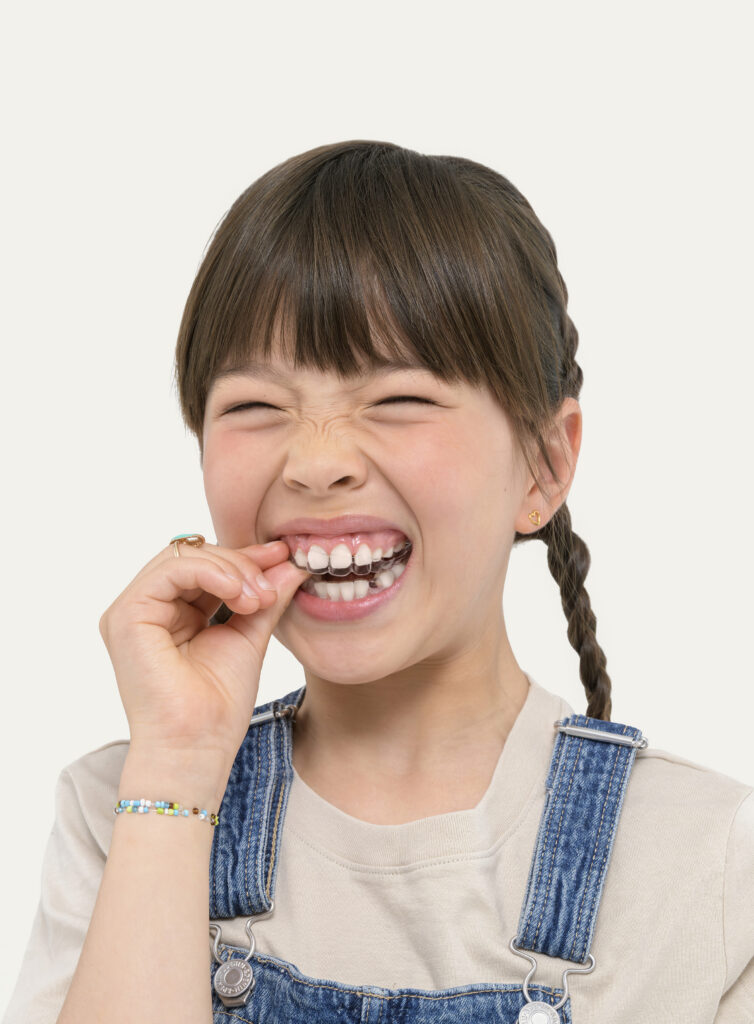 A young girl with braided pigtails winces playfully with squeezed-shut eyes as she fits a clear plastic orthodontic retainer over her upper teeth. She is wearing denim overalls over a beige shirt.