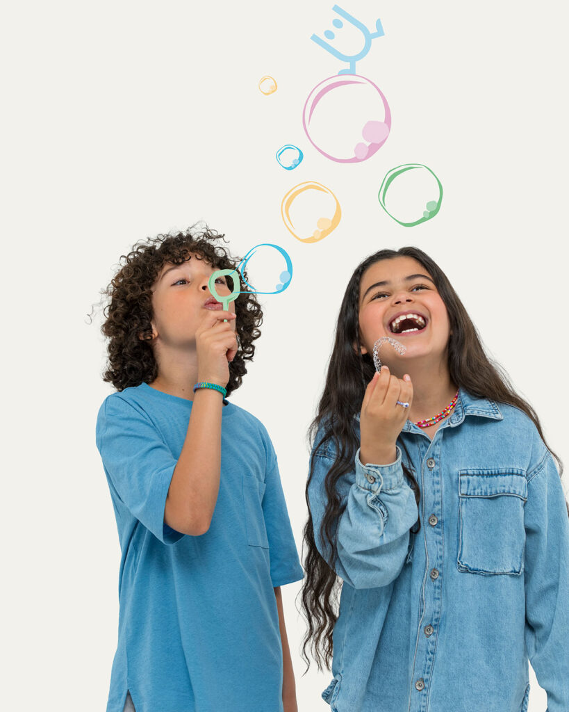 A young boy with curly hair in a blue T-shirt blows bubbles using a green wand, while a young girl with long dark hair in a denim shirt stands next to him, laughing joyfully. Playful, colorful hand-drawn bubbles float above them against a plain light grey background.