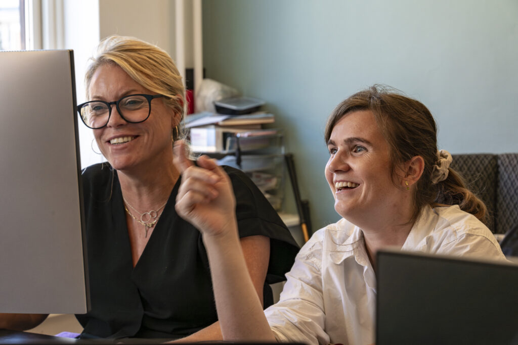 Two women sitting side-by-side at an office desk, smiling and looking at a computer screen together.
