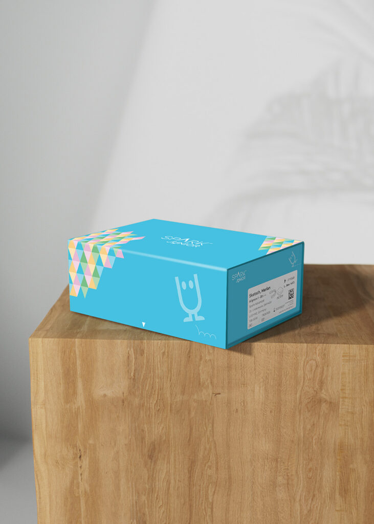 A rectangular bright blue 'SPARK JUNIOR' product box sitting on a wooden block against a plain wall. The box features a white cartoon character doodle on the side, a multicolored geometric triangle pattern on the corner, and a white product label on the right side.