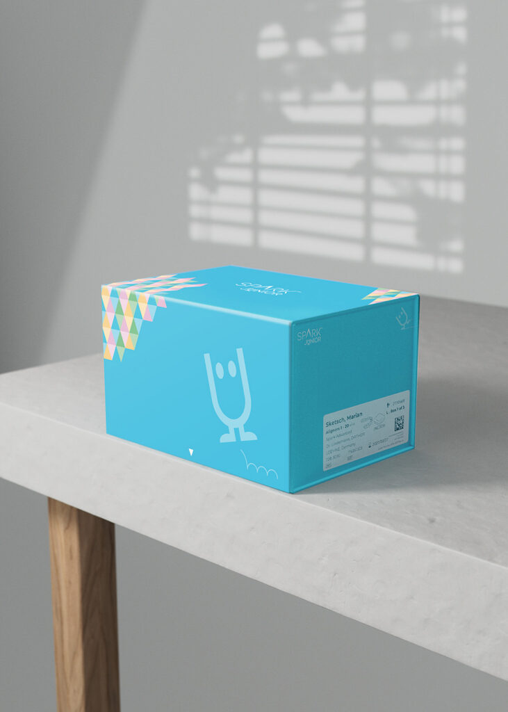 A bright blue 'SPARK JUNIOR' product packaging box displayed on a light concrete table. A shadow from a window blind falls across the white wall behind it, highlighting the box's colorful geometric pattern and white cartoon character design.