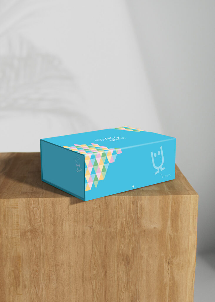 A bright blue 'SPARK JUNIOR' box shown from a slightly different angle on a wooden surface. The front displays a playful white cartoon character doodle, while the corner is covered in a colorful mosaic of pink, yellow, teal, and green triangles.