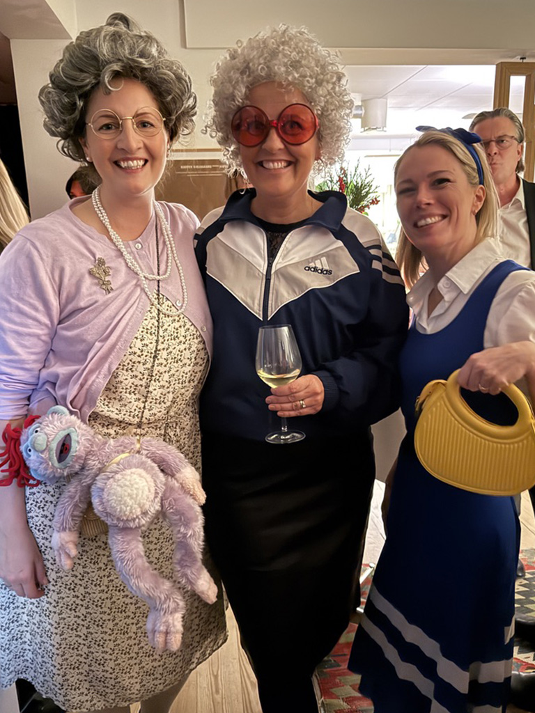 Three women dressed in humorous costumes at a party; two wear grey wigs and large glasses, while the third wears a blue dress with a yellow purse.