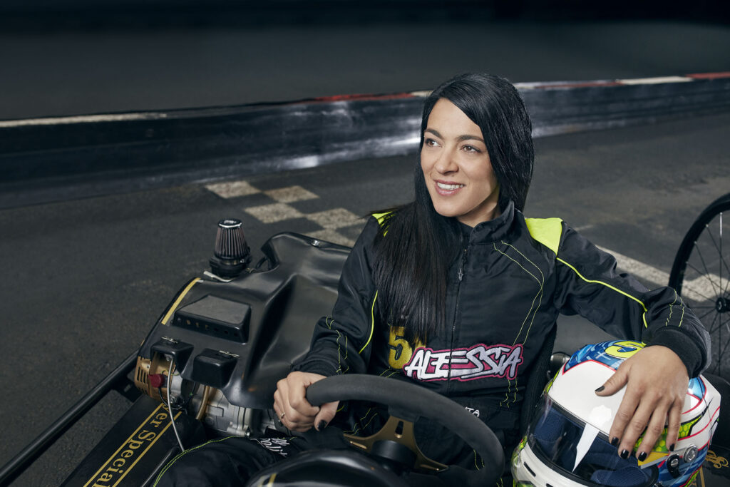 A dark-haired woman in a black and neon yellow racing suit sits in a go-kart on a track, smiling to the side while holding a white and colorful helmet in her lap.