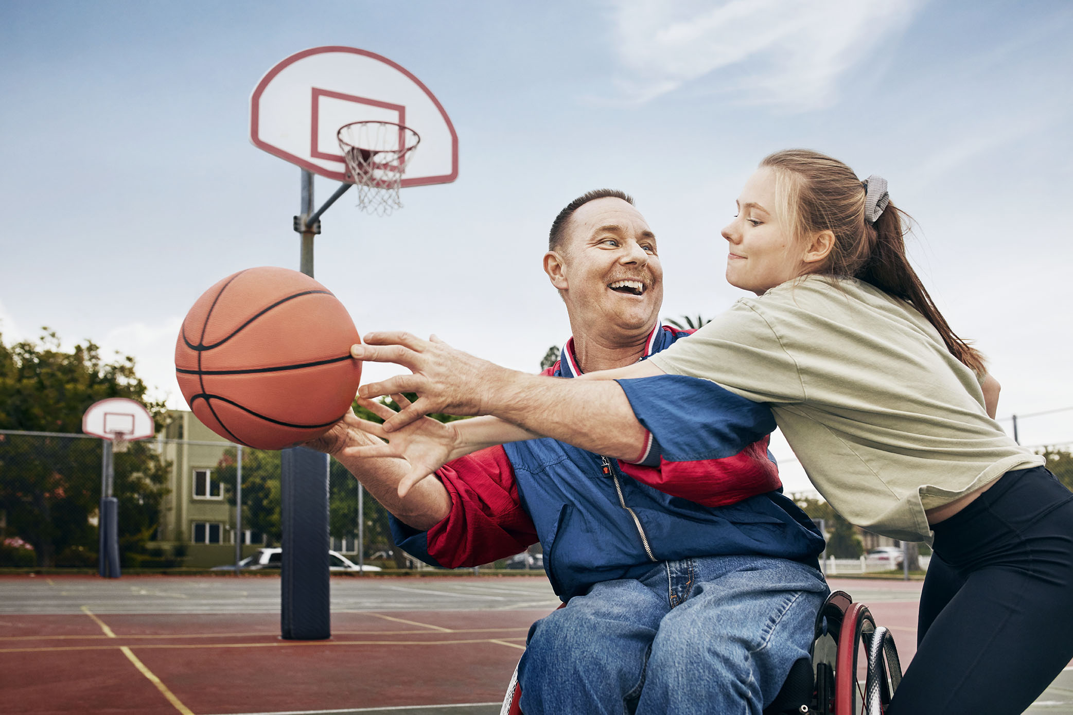 A man in a wheelchair and a young woman play basketball together on an outdoor court. Both are reaching up and smiling for a basketball in mid-air with the hoop in the background.