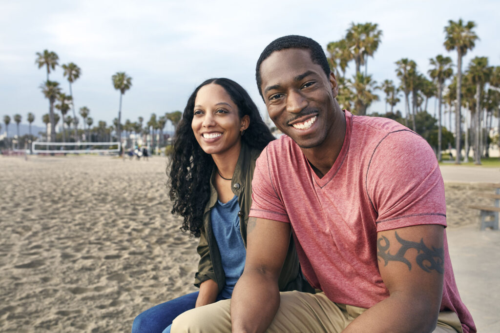 A smiling couple sits close together on a sandy beach with palm trees and a volleyball net in the background. The man wears a red T-shirt and has a tattoo on his upper arm.