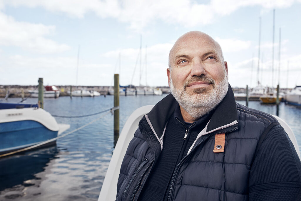A close-up of a smiling, middle-aged bald man with a grey beard looking upward. He is standing on a boat in a marina with masts and water in the background.