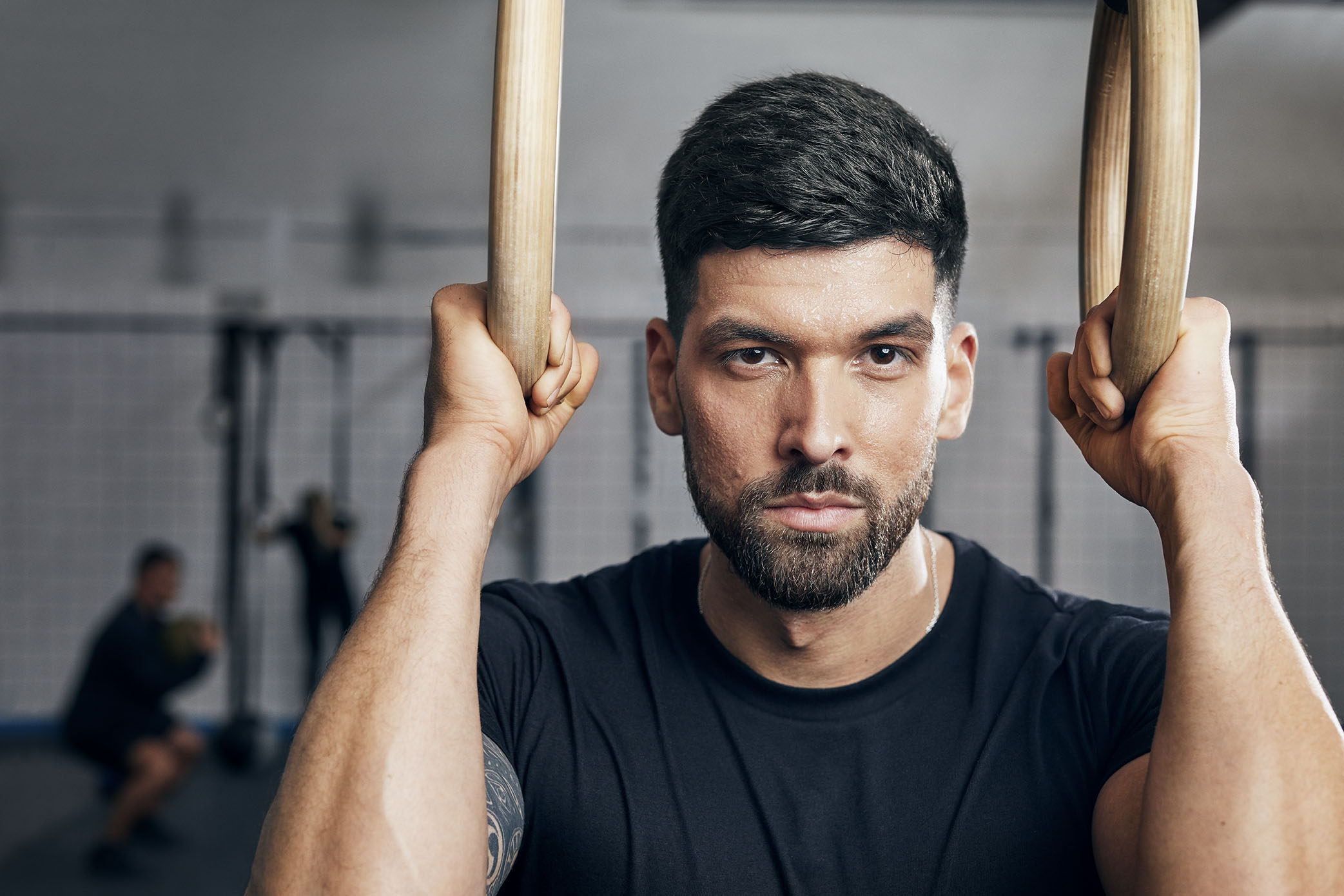 A portrait of a sweaty man with a beard looking directly into the camera with an intense gaze while holding onto two gymnastic rings in a gym.