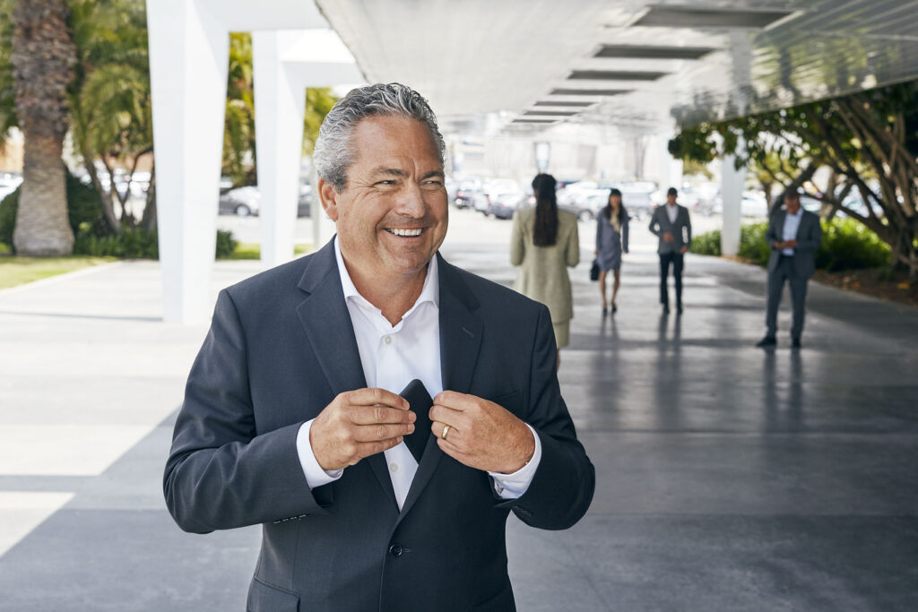 A smiling, grey-haired man in a dark suit and white shirt slides an object into his inside jacket pocket. The background shows a modern, open hallway with blurred people walking by.