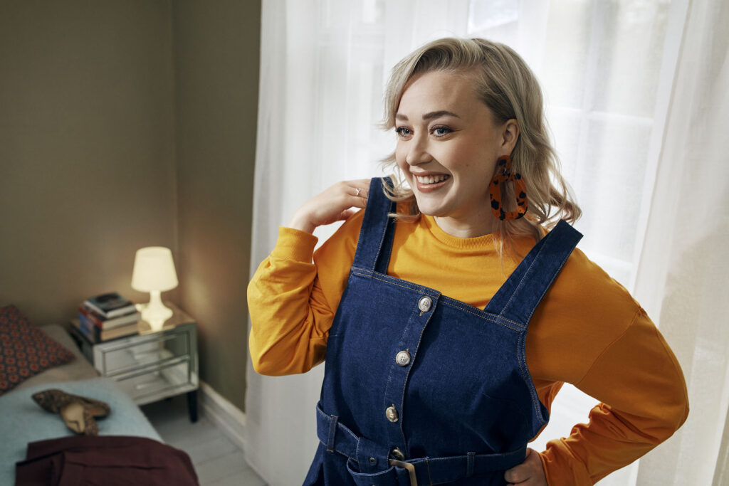 A smiling, blonde woman wearing a yellow long-sleeved shirt and dark blue denim overalls looks to the side while standing in front of a bright window.