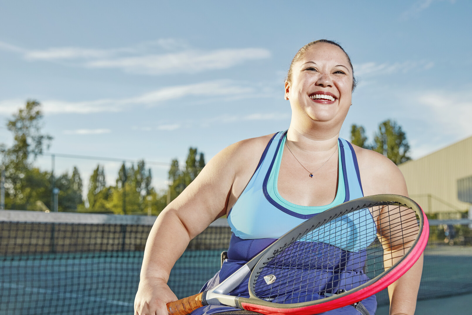 A smiling woman in a wheelchair on an outdoor tennis court under a blue sky. She is wearing a light blue sports top and holding a red and grey tennis racket in her lap.