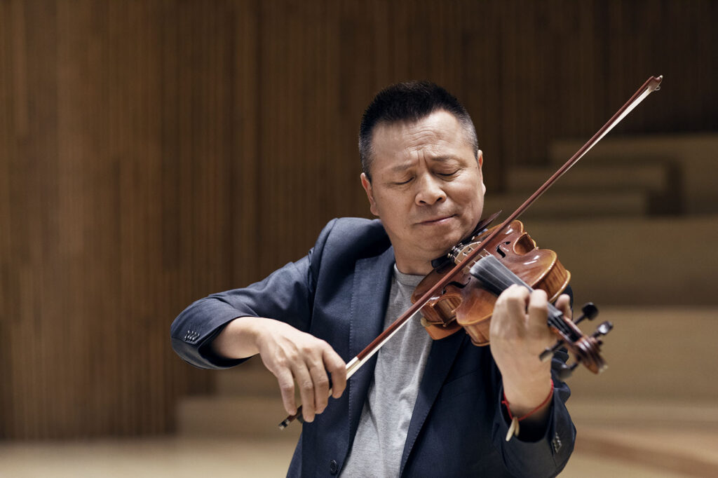 An Asian man in a suit plays the violin with deep concentration and closed eyes inside a concert hall with wooden panels in the background.